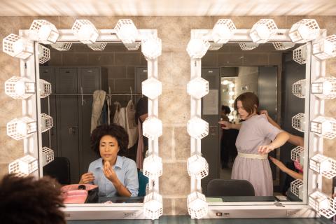 Two people are in a dressing room with two large mirrors framed by bright lights. The image captures a behind-the-scenes moment, likely in a theater or performance setting, highlighting the preparation process.